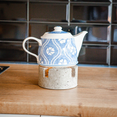 Auf einer Arbeitsplatte in einer Küche steht ein Stövchen aus Beton auf der eine Teekanne mit blauen Blumen steht. In dem Stövchen stehen Teelichter. 