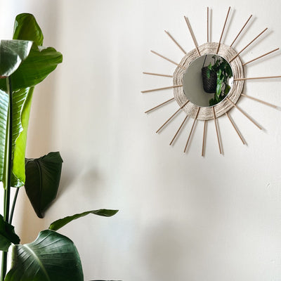 Ein runder Spiegel mit einem sonnenförmigen Holzrand hängt an einer weißen Wand. Daneben ist eine große Pflanze sichtbar. 
